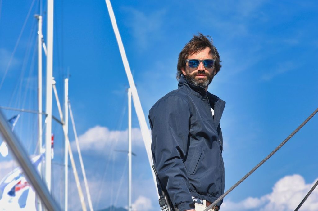 Bearded man in a navy jacket and sunglasses on a sailboat, illustrating staying active while monitoring TRT side effects